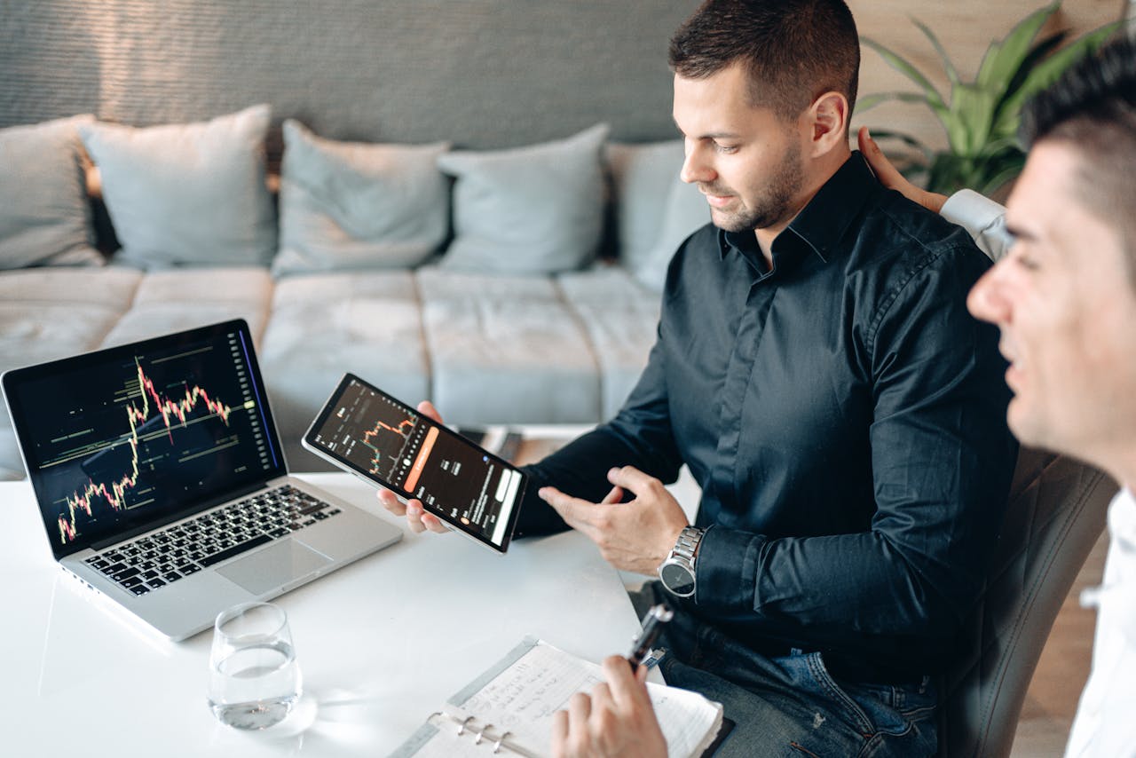 Two businessmen discussing financial data on a tablet and laptop, analyzing line graphs in a modern office setting.